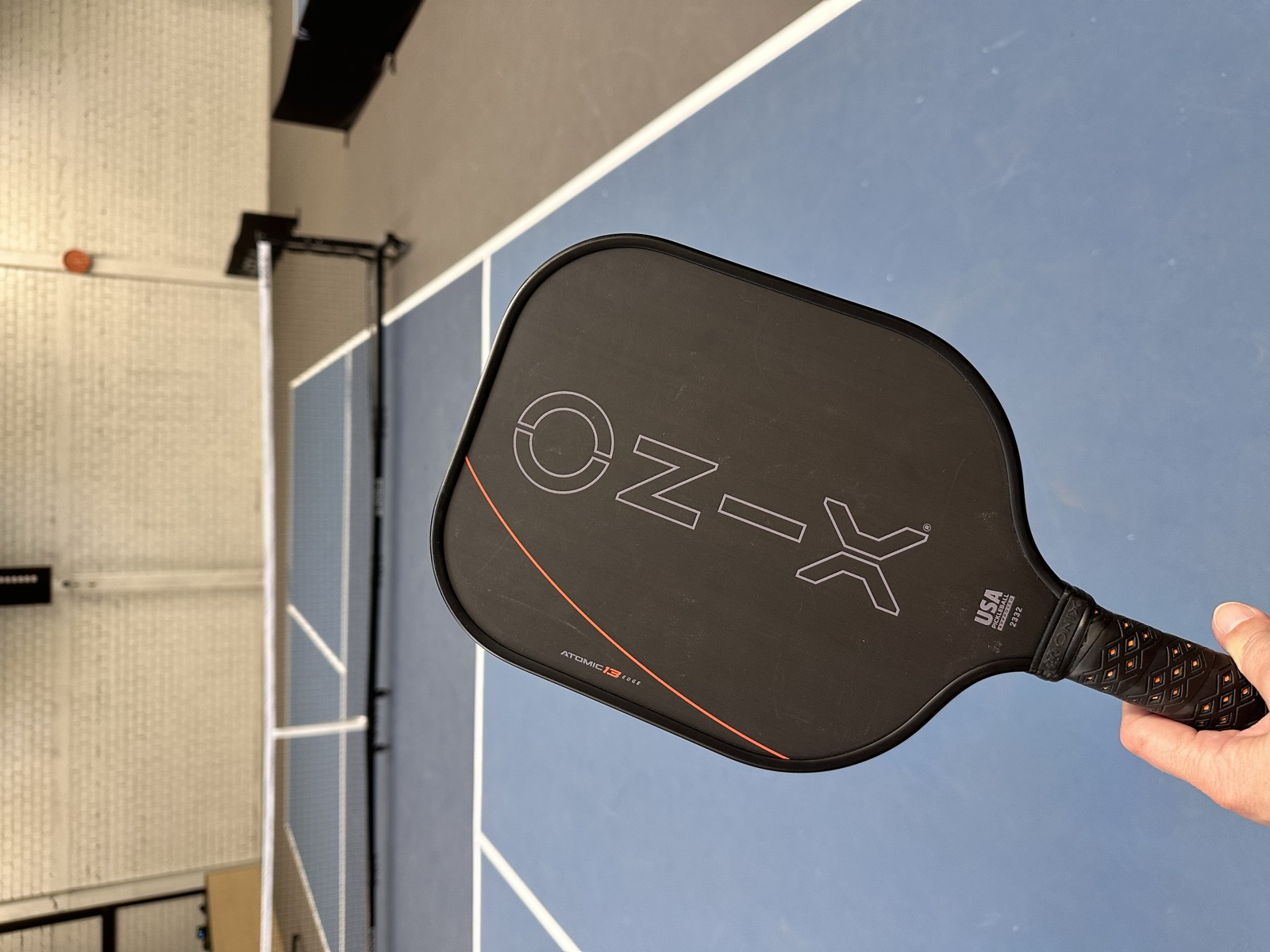 Onix Evoke Premier Raw Carbon 16 mm in der Hand auf einem Indoor-Pickleball-Court